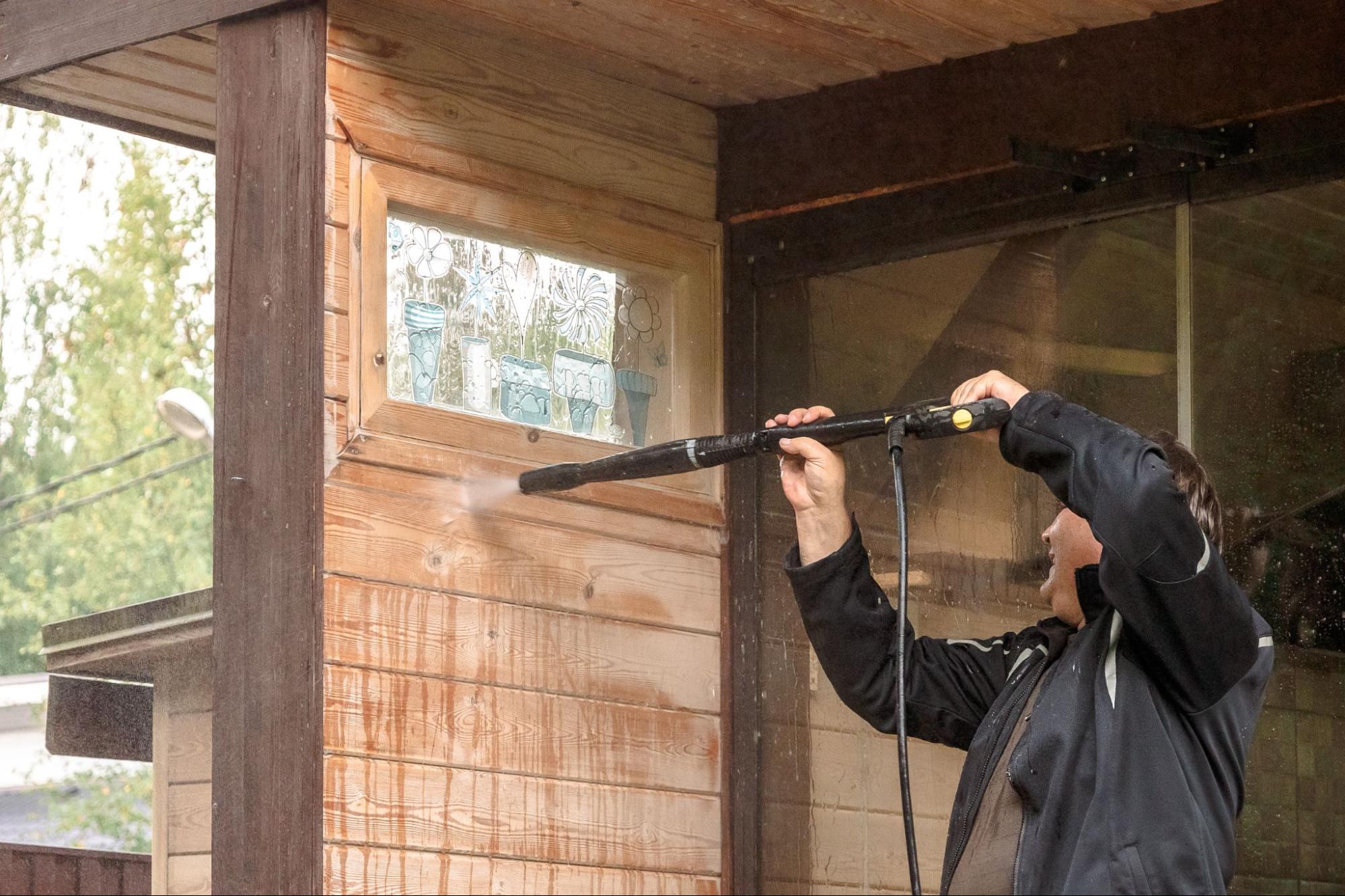 A man pressure washes exterior wood planks below a stained glass window.