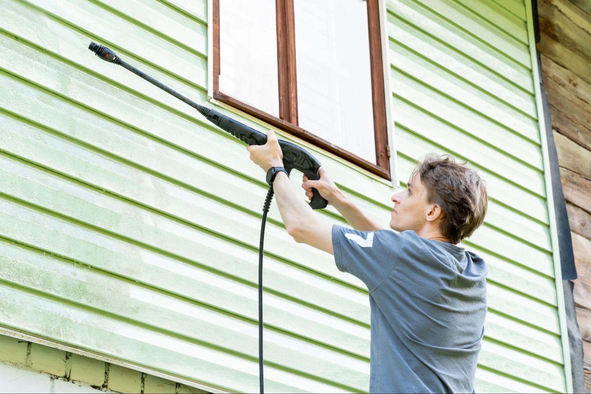 A man uses a pressure washer to clean the green face of his home.