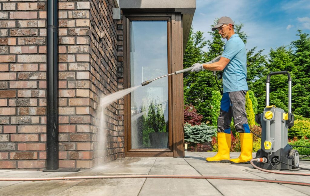 A man in yellow rain boots pressure washing brick facing on a home.
