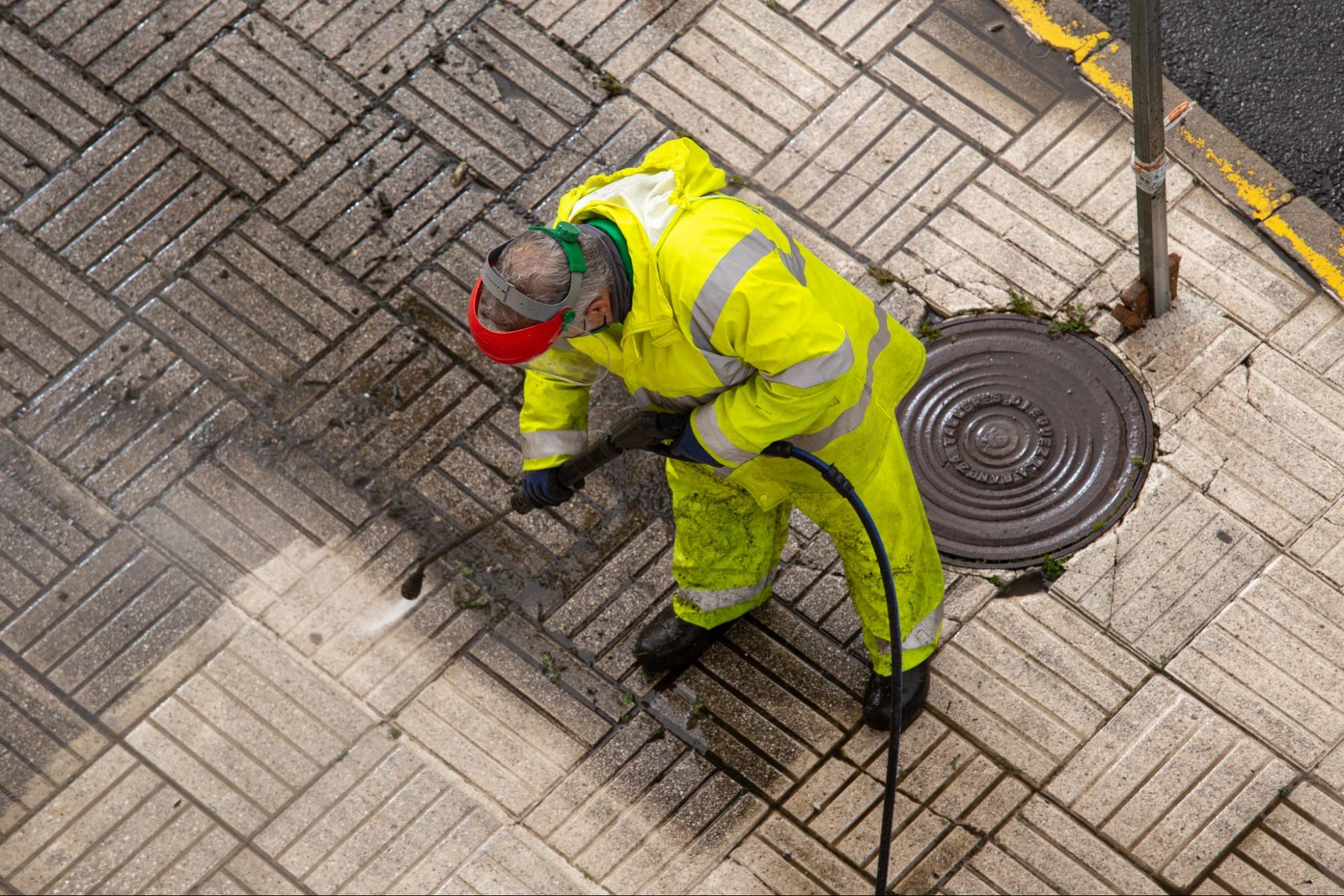 Top view of a worker in a fluorescent uniform, pressure washing a street sidewalk