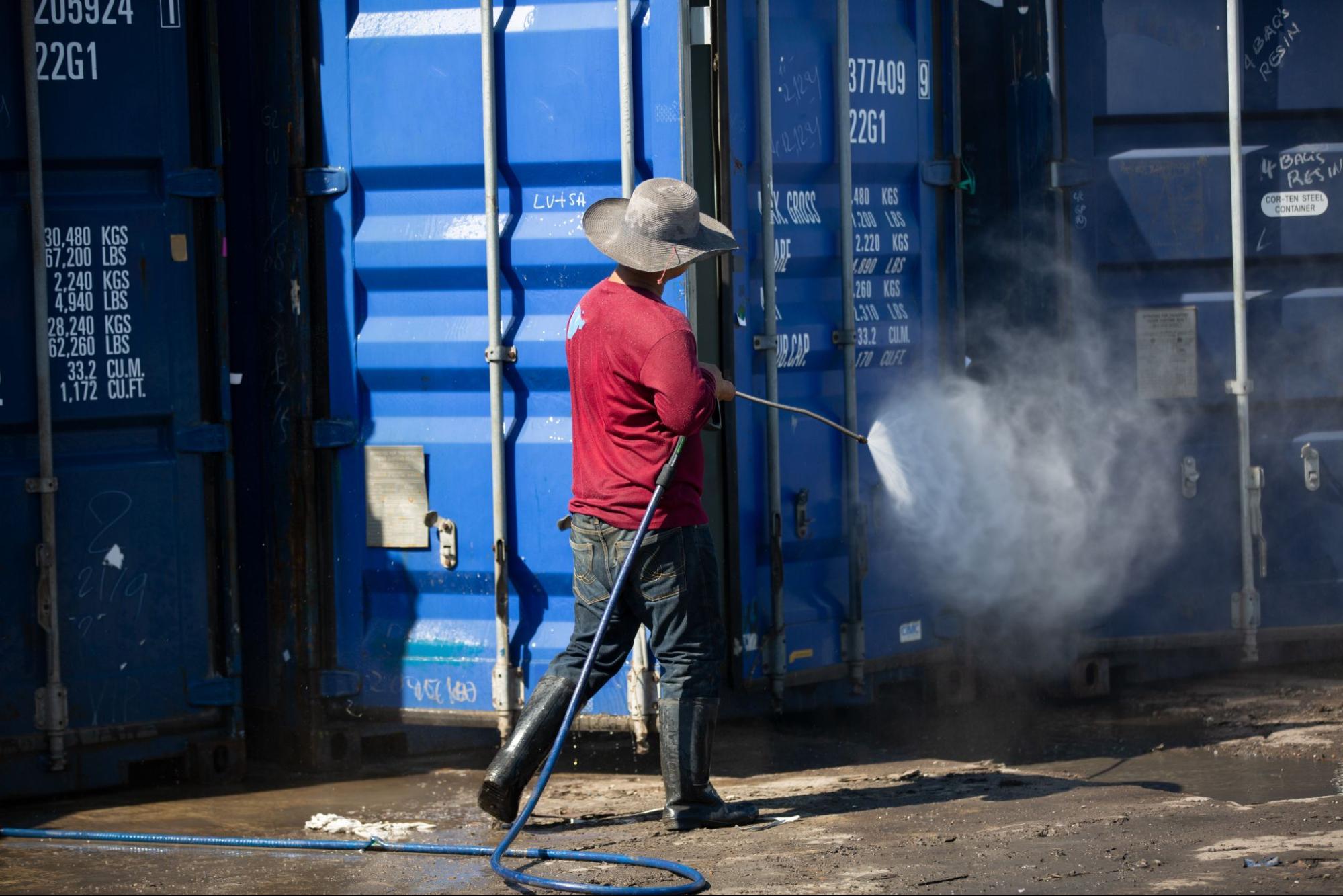 A worker cleans the cargo containers at a port shipyard using an industrial pressure washer