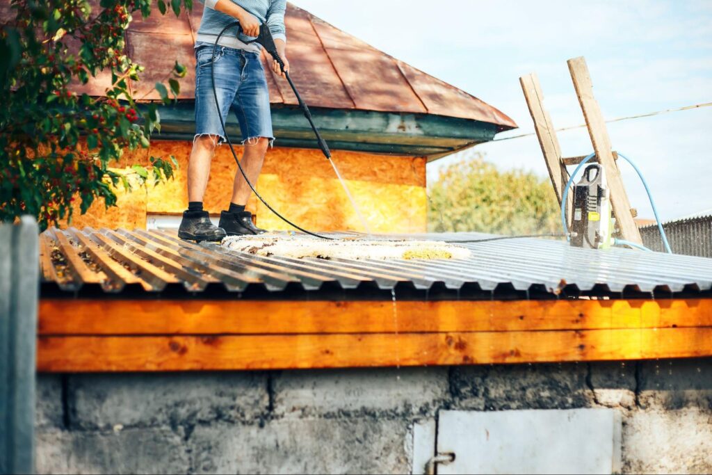A man in jean shorts stands on a roof and pressure washes it.